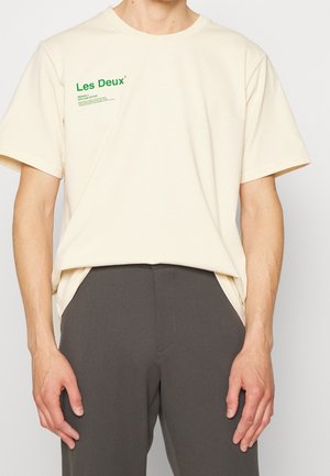 Man draagt een crèmekleurig Les Deux T-shirt met groen logo op de borst, gecombineerd met donkergrijze broek tegen een effen achtergrond.