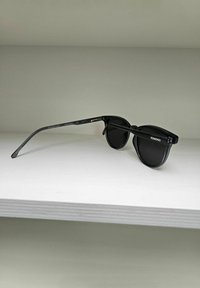 Lunettes de soleil noires en plastique avec des verres ronds et une finition brillante subtile. Les branches minces et courbées portent le nom de la marque "KOMONO" et une étiquette de protection UV.