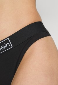 Bikini en coton noir avec une large ceinture élastique arborant un logo contrastant. Le tissu semble lisse avec une coupe ajustée.
