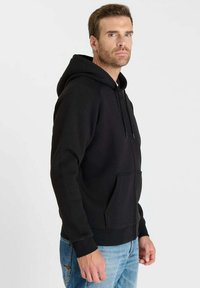 Zwarte hoodie met een voorzak, een capuchon met trekkoord en geribbelde manchetten. Draagt met lichtblauwe spijkerbroek. Eenvoudig ontwerp, gladde textuur.