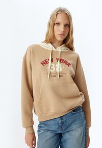 Sudadera beige con una capucha amarilla claro contrastante, que presenta un gráfico rojo "NUEVA YORK 38" y un bolsillo frontal. Corte clásico relajado.