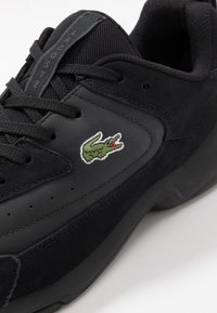 Czarny sportowy but wykonany ze skóry i siateczki, z teksturowaną czarną zamszową wstawką, sznurowanym zapięciem oraz zielonym logo krokodyla Lacoste.