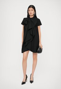 Vestido preto curto com um laço grande e dramático na gola, apresentando suaves pregas. Combinado com sapatos de salto altos pretos e uma pequena bolsa preta.