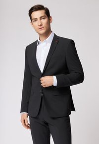 Suit elegante nero con rever a scapola e chiusura a un bottone. Camicia bianca sotto. Tessuto grigio scuro con una lieve trama.