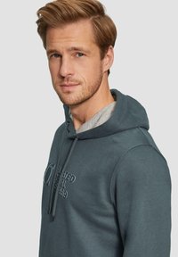 Sweatshirt à capuche en tissu bleu canard avec une texture lisse. Dispose d'un intérieur gris, d'un cordon de serrage réglable et de détails logo sur le devant.