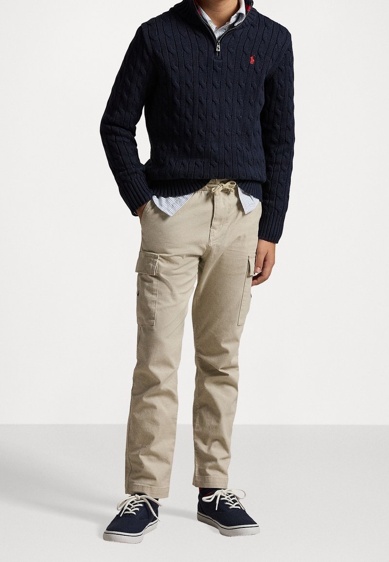 Navy cable-knit sweater with half-zip, beige cargo pants with pockets, and dark sneakers with white soles. Structured, casual outfit.