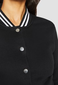 Černá varsity bunda s žebrovaným límcem, který má bílé pruhy a stříbrné cvoky, vyrobená z hladké látky. Detailní pohled na design.