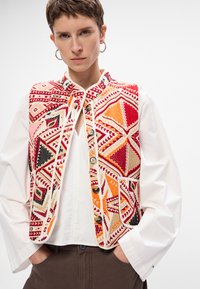 Színes hímzett mellény törzsi geometriai mintával piros, narancs és fekete színekben. Elülső gombos záródással és oldalsó zsebbel rendelkezik.