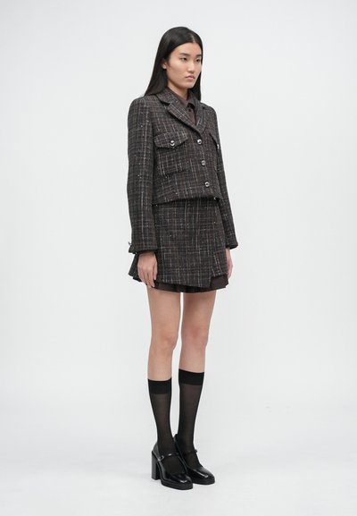 Pruun tweedist jakk kahe esikotiga, millele on lisatud sobiv seelik. Mudel kannab põlvikutega musta värvi sokke ning musti plokktaldkingi.