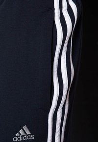 Detailní záběr na námořnické modré kalhoty Adidas se třemi bílými pruhy po stranách a vyšívaným logem poblíž kapsy.