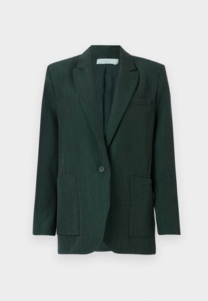 Henkilö pukeutunut tumman sävyiseen, oversized bleiseriin, harmaaseen hameeseen, jossa valkoiset pitsireunat, mustiin korkokenkiin ja pitää mustaa nahkalaukkua.