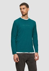 Turkusowa koszula z długim rękawem o teksturowanej powierzchni i kontrastującym białym wewnętrznym kołnierzyku. Na przodzie umieszczony jest wytłoczony napis "PRZESTRZEŃ JEST MIEJSCEM".