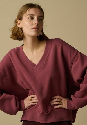 Jeune femme aux cheveux courts portant un pull marron ample à encolure en V, plusieurs bagues en or, ainsi que des boucles d'oreilles créoles et clous en or, sur un fond neutre.