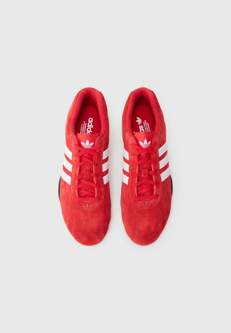 Chaussures de sport en daim rouge avec des bandes en cuir blanc, bout rond, lacets et semelle en caoutchouc noire. L'intérieur présente un logo et une marque.