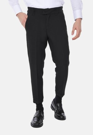 Uomo che indossa pantaloni neri su misura, calzini neri e mocassini neri con nappine, con una mano in tasca e una camicia bianca.