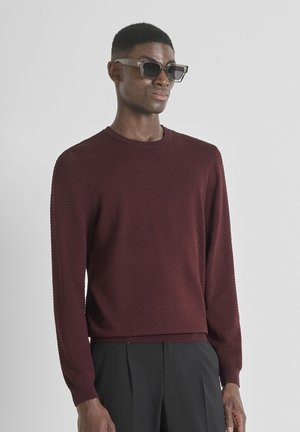 Maglione bordeaux texturizzato con maniche lunghe e colletto rotondo, abbinato a occhiali da sole rettangolari. Il tessuto presenta un motivo sottile.