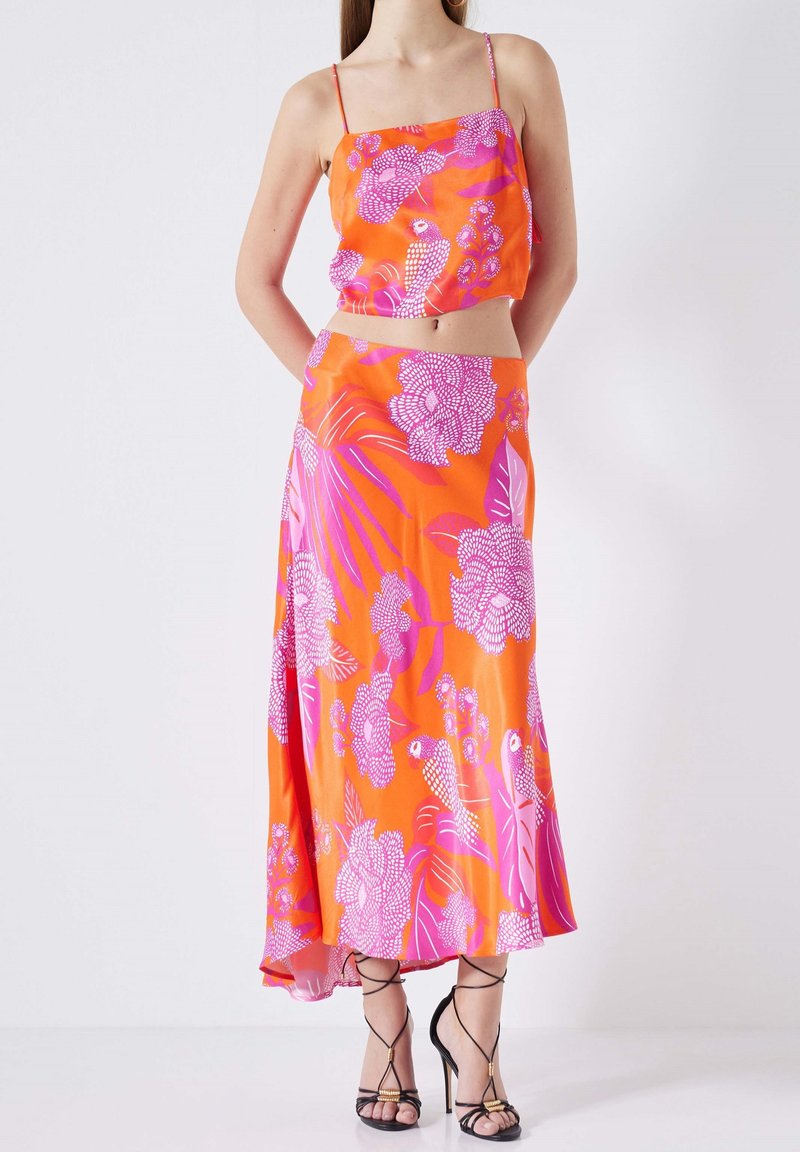 Femme portant un crop top à fines bretelles imprimé de fleurs orange et rose, assorti à une jupe midi taille haute, accompagnée de talons noirs à lanières.