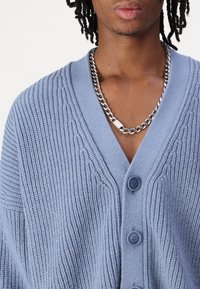 Ljusblå stickad cardigan med V-ringning, med ribbad textur och tre knappar fram, kompletterad med ett silverfärgat kedjearmband.