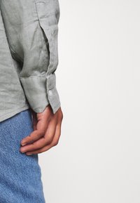 Camisa de lino verde claro con puños abotonados, presenta una textura visible y un corte relajado, combinada con tejido de denim azul.