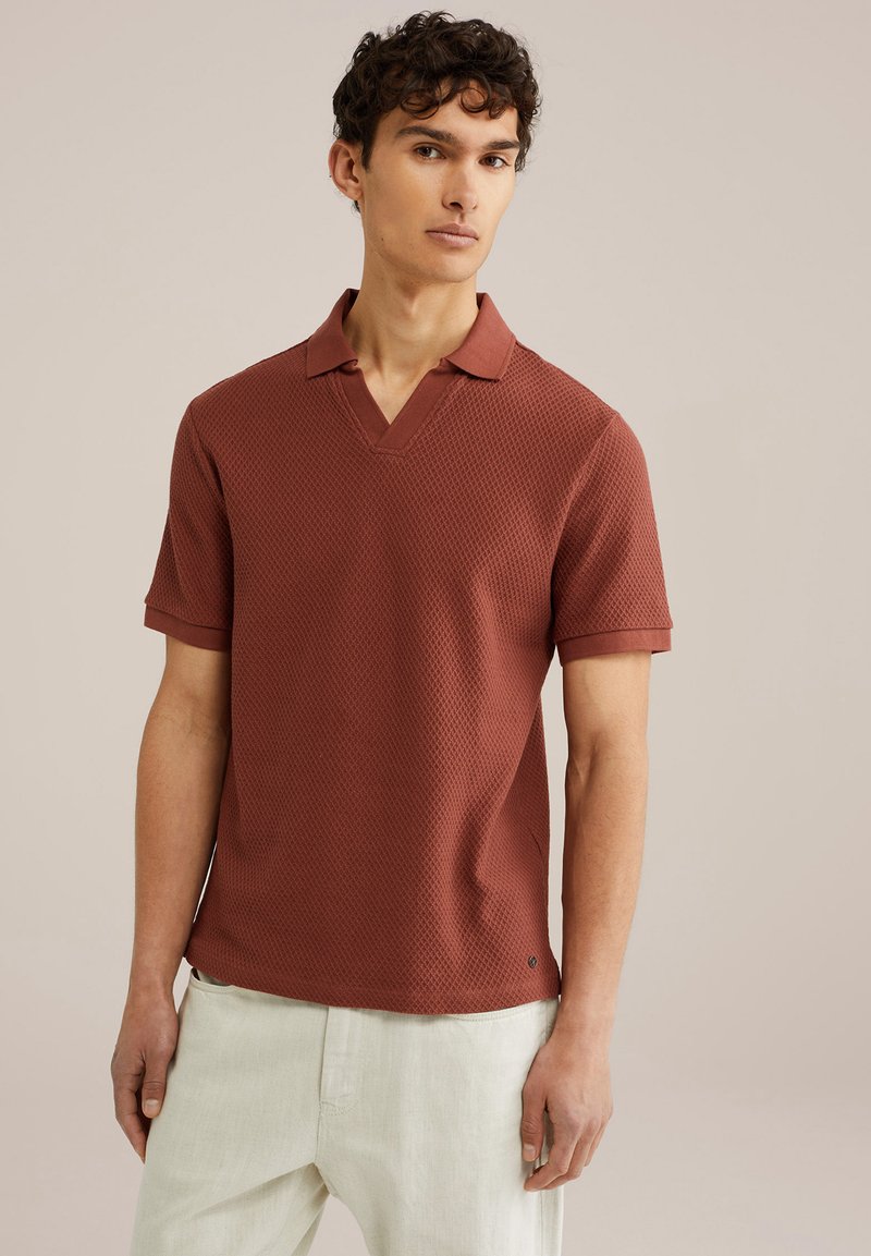 Rust-colored short-sleeve polo shirt with a textured pattern, V-neckline, and solid collar; paired with light-colored trousers.