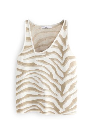 Femme portant un pantalon flare blanc, un haut sans manches beige et blanc à motifs, et des sandales à talons hauts dorées, debout contre un mur rose.