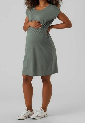Robe de maternité en tissu doux vert olive, manches courtes, taille froncée et coupe décontractée ; associée à des baskets en toile blanches.