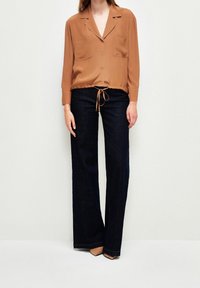 Bruine, lange mouwen, zijden button-up shirt met een trekkoord onderkant, gecombineerd met donkere spijkerbroek met wijde pijpen en beige hakken.