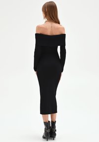 Schwarzes, geripptes, gestricktes Off-Shoulder-Kleid mit langen Ärmeln, figurbetonter Silhouette und mittlerer Wadenlänge. Kombiniert mit schwarzen Stiefeletten mit dekorativen Metallakzenten.