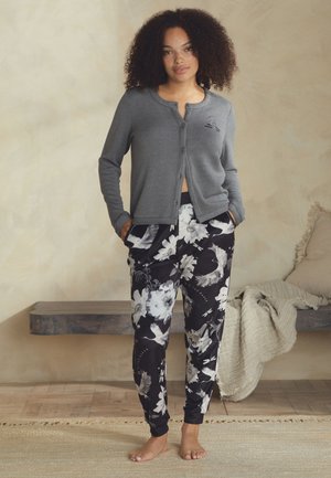 Woman stands barefoot in gray button-up cardigan and black floral print pants in a minimalist room with beige walls and wooden floor.