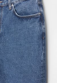 Gros plan sur un jean en denim bleu montrant la poche avant, la boucle de ceinture et les coutures orange sur un tissu texturé.