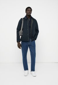 Casual dark blue jacket over a navy shirt, paired with light blue jeans and white sneakers. Gray shoulder bag and textured fabric details visible.