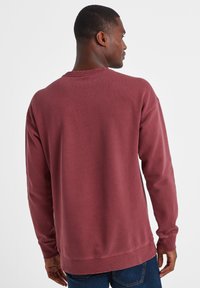 Man wearing a long-sleeve, crew-neck maroon sweatshirt and blue jeans, seen from behind, facing slightly to the right.