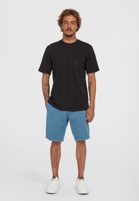 Zwart T-shirt met korte mouwen en een borstzak, gecombineerd met lichtblauwe shorts. Model draagt witte sneakers. Eenvoudig, casual ontwerp.