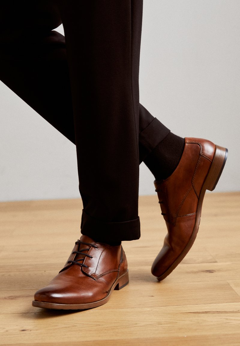 Braune Leder Schnürschuhe mit glatter Oberfläche, runder Zehenpartie und niedrigem Absatz, kombiniert mit hochgerollten schwarzen Hosen und Socken.