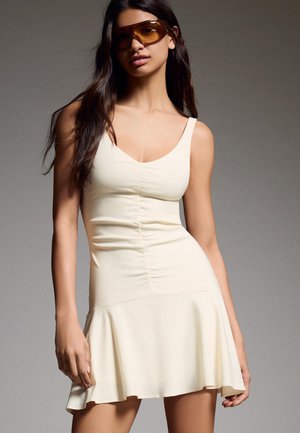 Donna con lunghi capelli scuri che indossa grandi occhiali da sole marroni e un vestito aderente senza maniche color crema con orlo svasato.