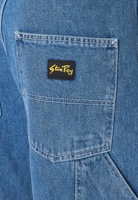 Denim nadrág közeli felvétele kék anyaggal, fekete címkés zsebbel, fehér varrással és texturált felülettel.