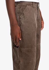 Brown cargo pants made of textured fabric, featuring side pockets, visible stitching, and a relaxed fit, shown from a side angle.