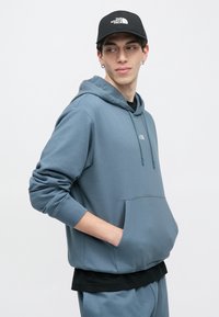 Ung mann iført en blå hettegenser og joggebukse med hendene i frontlommen, ser sidelengs, og har også på seg en svart caps med hvit logo.