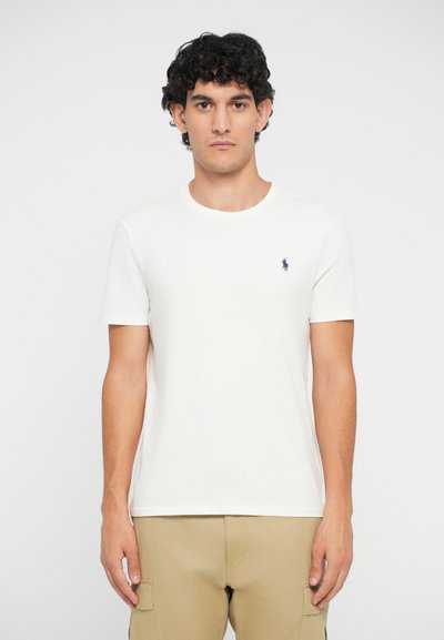 Moški s kodrasto temno lasmi, oblečen v enobarvno belo majico s kratkimi rokavi, ki ima majhen modri izvezeni logotip, in bež cargo hlačami.