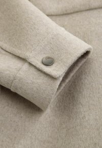 Manche d'un manteau en laine beige avec un seul bouton pression en métal brillant sur le poignet, mettant en valeur la texture du tissu et les détails de la couture.