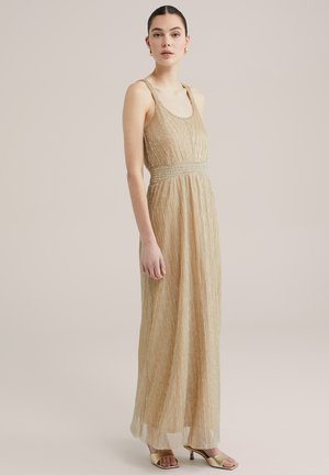 Robe maxi plissée dorée avec des bretelles réglables, taille smockée et ourlet transparent. Modèle associé à des talons ouverts dorés.