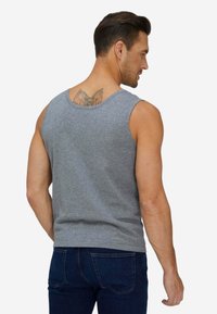 Uomo che indossa una canotta grigia senza maniche e jeans blu scuro, mostra un tatuaggio sulla parte superiore della schiena, in piedi contro uno sfondo chiaro uniforme.