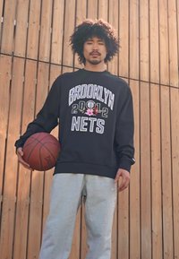 Felpa nera con grafica "BROOKLYN 2012 NETS", abbinata a pantaloni della tuta grigi. Tiene un pallone da basket, posizionandosi contro uno sfondo di legno.