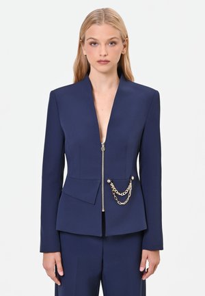 Giovane donna con capelli biondi lunghi che indossa un blazer blu navy con zip frontale e catena dorata decorativa sulla tasca.