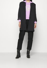 Cappotto nero sopra una felpa lilla con logo, abbinato a pantaloni neri e stivali con tacco neri. Il tessuto appare liscio e strutturato.