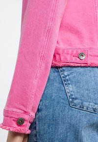 Chaqueta de mezclilla rosa con dobladillo deshilachado, detalle de botones en las mangas y un cuello clásico, combinada con pantalones cortos de mezclilla azul.