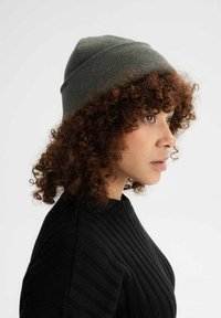 Grijze gebreide beanie met een taps toelopende top en ribbelstructuur. Heeft een strakke pasvorm en wordt gedragen met een zwarte ribbeltrui.