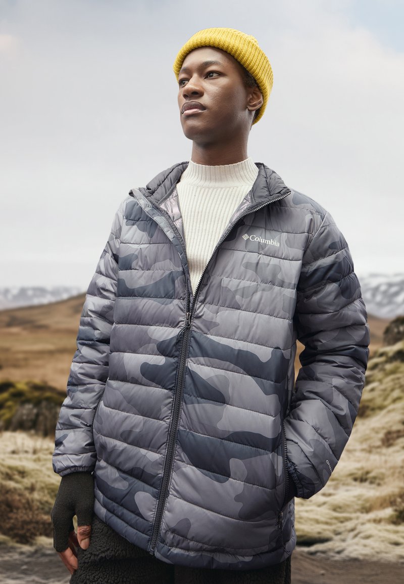 Jeune personne portant une veste Columbia grise à motif camouflage, un bonnet jaune en laine et des gants noirs sans doigts dans un paysage montagneux extérieur.