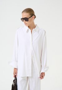 Valkoinen button-up-paita, joka on valmistettu pehmeästä kankaasta, pitkähihaisena ja klassisella kauluksella. Malli pitää mustaa käsilaukkua ja käyttää tummia aurinkolaseja.
