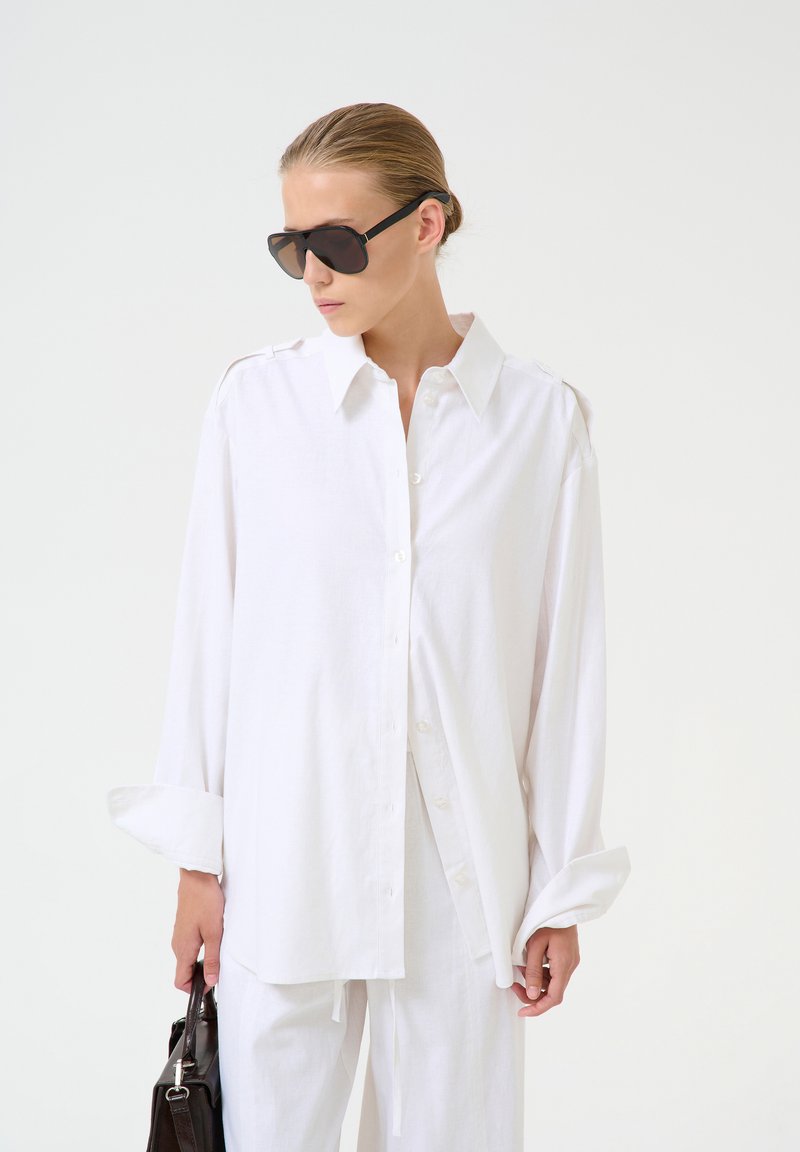 Valkoinen button-up-paita, joka on valmistettu pehmeästä kankaasta, pitkähihaisena ja klassisella kauluksella. Malli pitää mustaa käsilaukkua ja käyttää tummia aurinkolaseja.